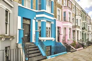 A row of colorful terraced houses with distinct architectural styles, including a blue house with a black door and white trim, a pink house with a white door, and a green house with a white door. - Stylish Notting Hill Apartment (Greater London)