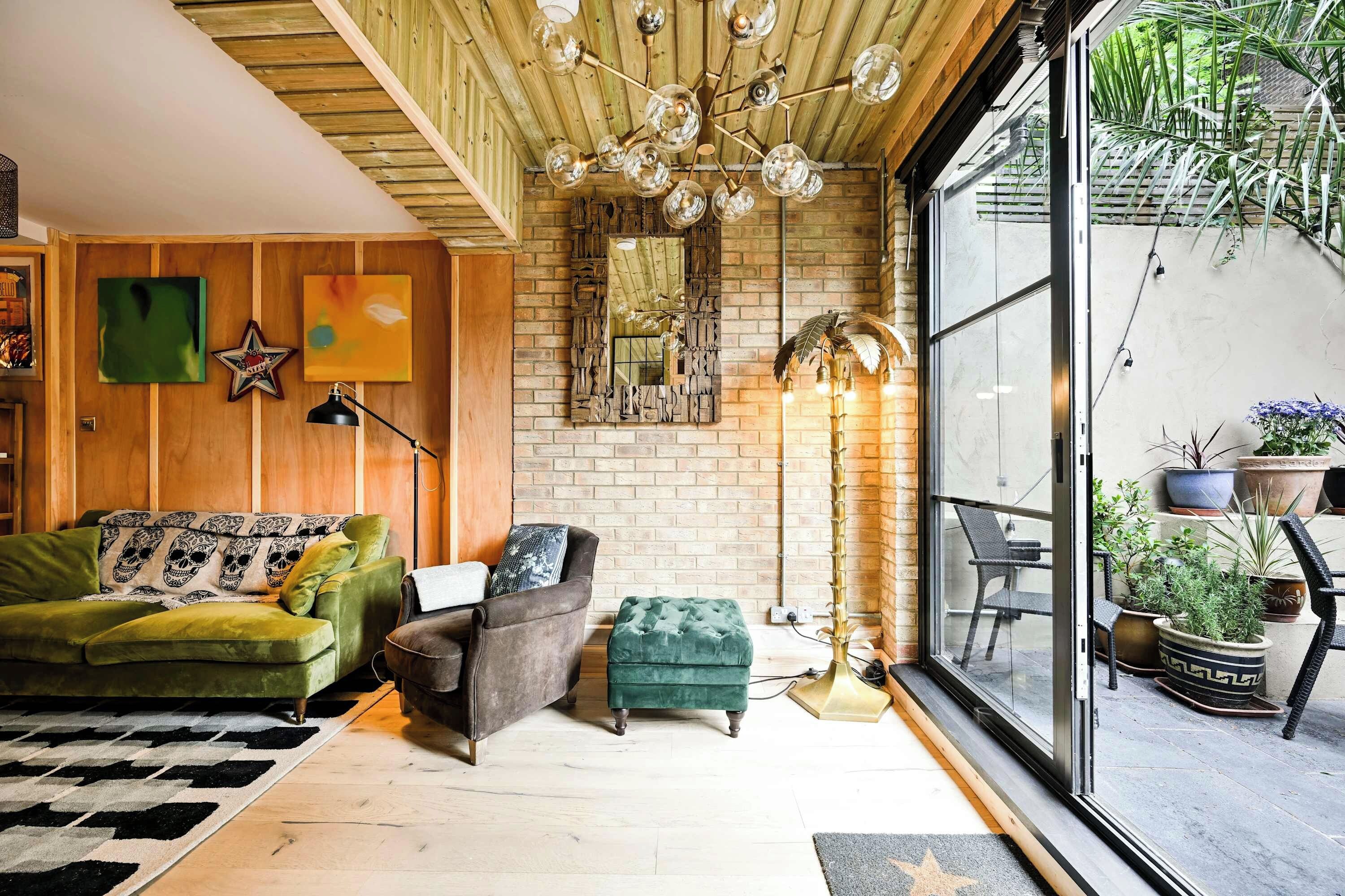 A cozy living room with a green sofa, a brown armchair, and a green ottoman. There is a large mirror on the wall, a chandelier, and potted plants outside the window.