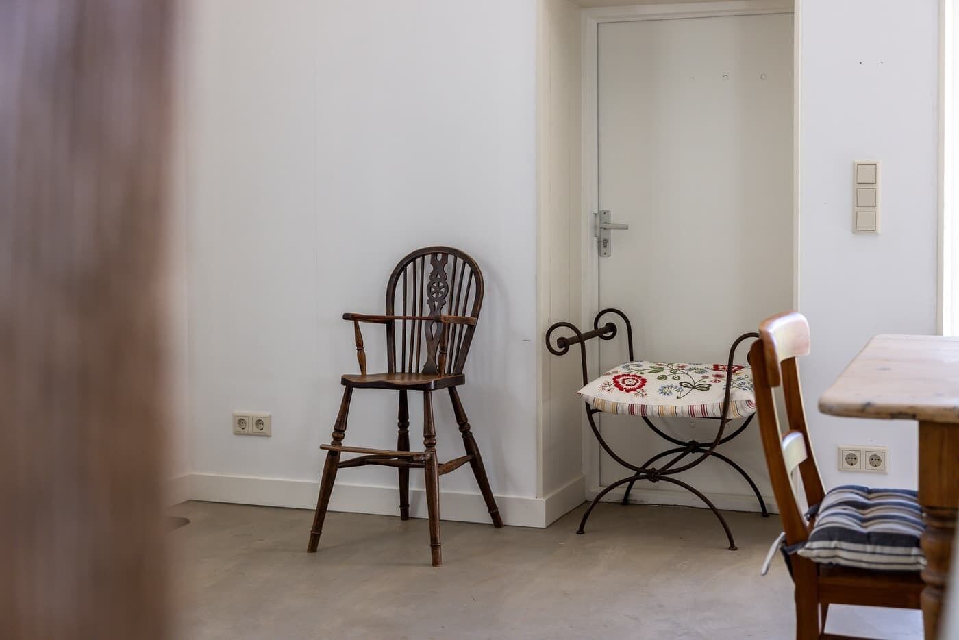 A room with a wooden chair, a metal chair with a floral tablecloth, and a white door.