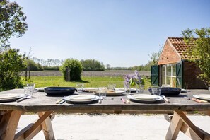 A wooden table set for dining outdoors with a view of a field and a small brick building in the background. - Holiday home for 8 in Oostkapelle | Oranjezonweg 7 - Klein Overduin (Oostkapelle)