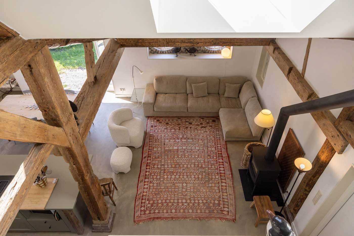 Et rum under taget med synlige træbjælker, en sofa, et mønstret tæppe, en stol og et skrivebord med en lampe.