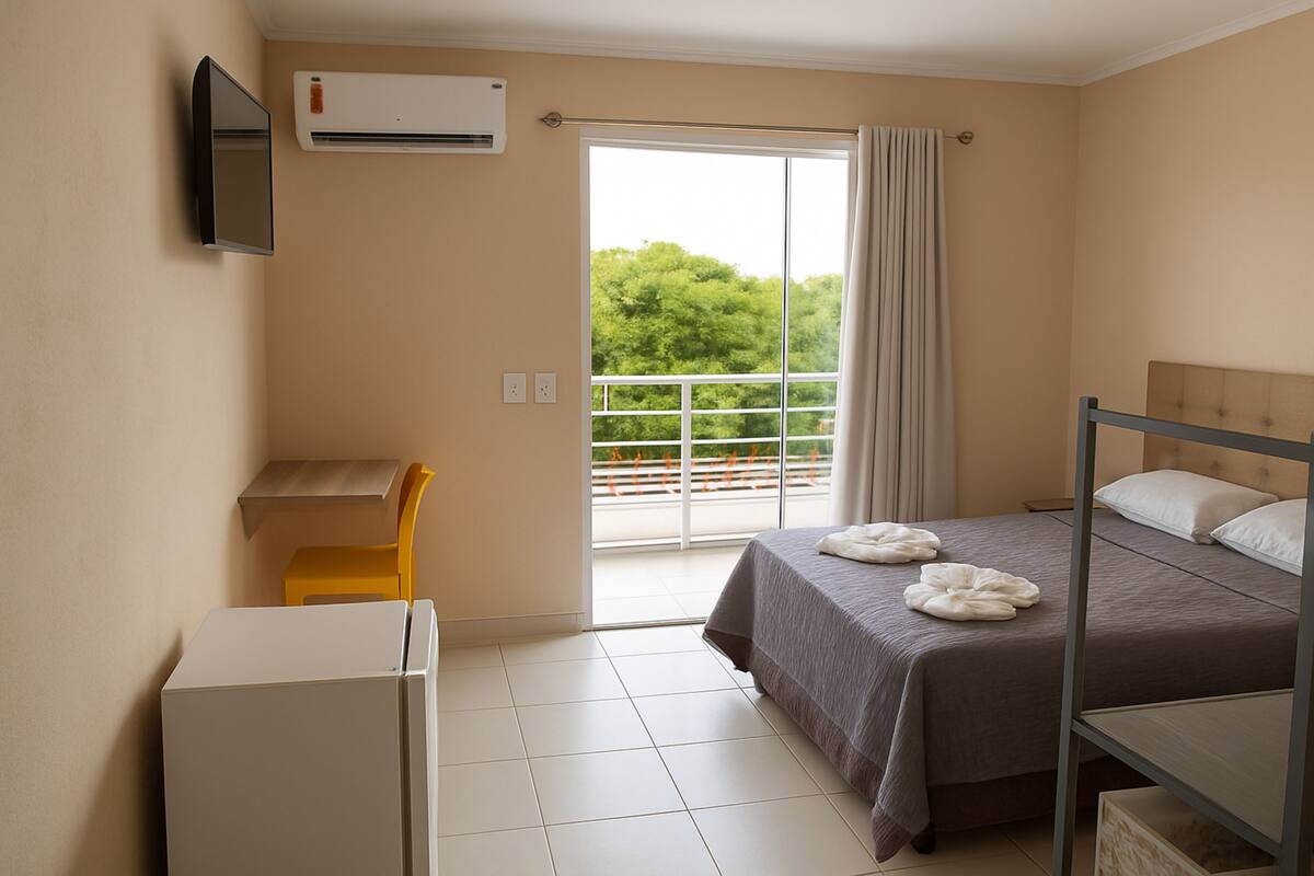 Quarto de hotel com cama, escrivaninha, cadeira, ar condicionado e vista para o verde pela janela.