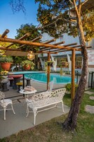 Uma área de piscina com pérgola, móveis de vime branco e plantas em vasos.