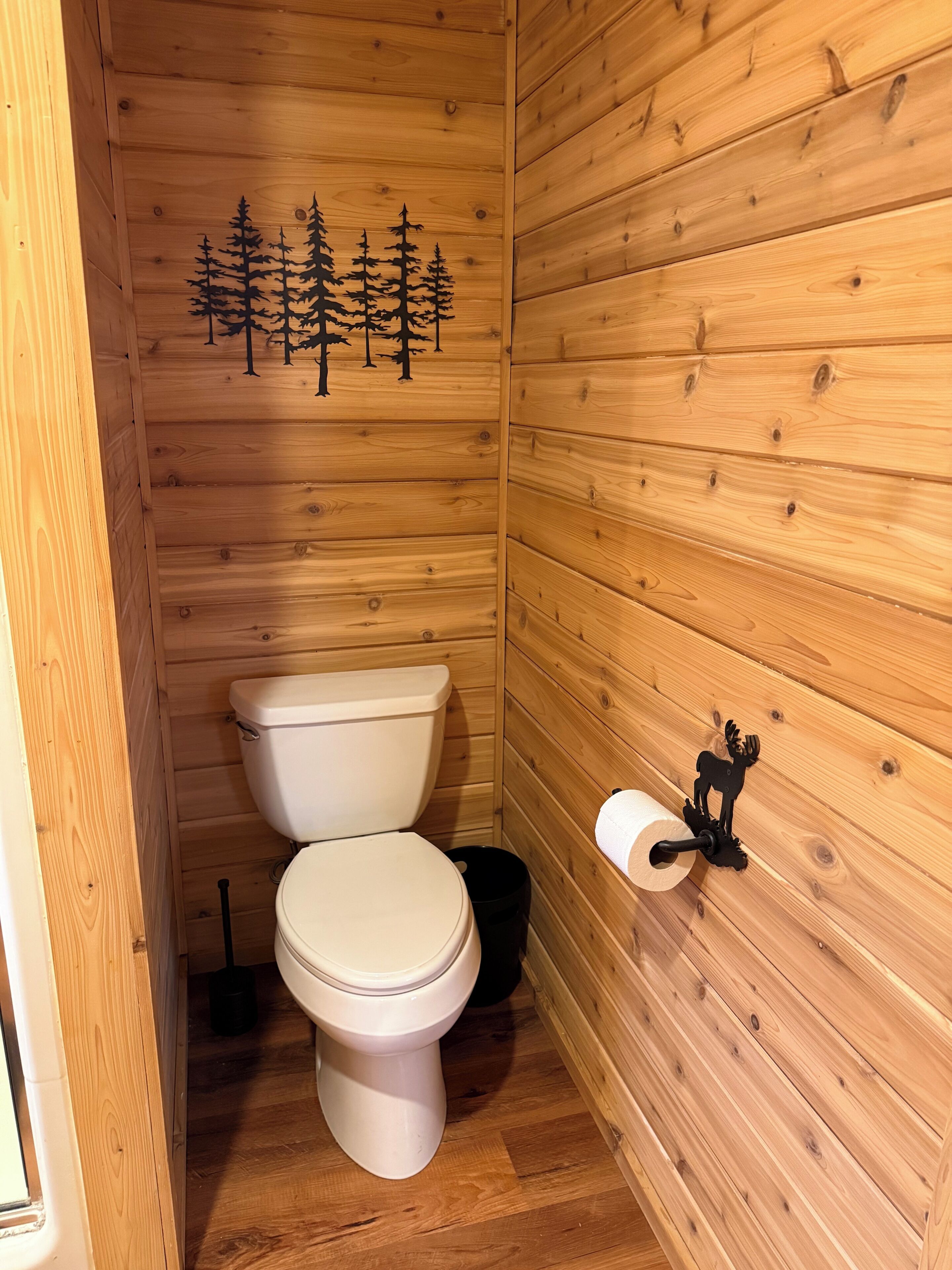 Une salle de bain aux murs lambrissés, équipée de toilettes, d’une poubelle noire et d’un porte-rouleau de papier toilette orné d’une décoration représentant un cerf.