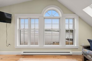A room with a large bay window overlooking a beach, a flat-screen TV mounted on the wall, and a built-in heater.