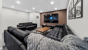 A modern living room with a large flat-screen TV, black leather sofas, a wooden coffee table, and framed pictures on the wall.