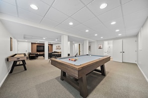 A pool table in a well-lit room with a carpeted floor, a foosball table, and a seating area with a TV.