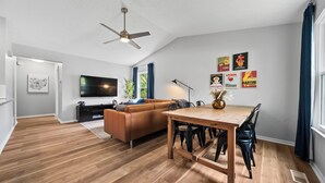A modern living room with a wooden floor, a brown sofa, a dining table with chairs, a ceiling fan, and a TV mounted on the wall.
