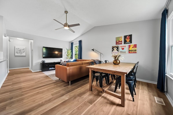 A modern living room with a wooden floor, a brown sofa, a dining table with chairs, a ceiling fan, and a TV mounted on the wall.