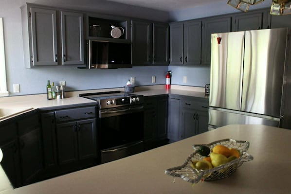 A kitchen with grey cabinets, a stainless steel refrigerator, a stove, and a fruit basket on the counter.