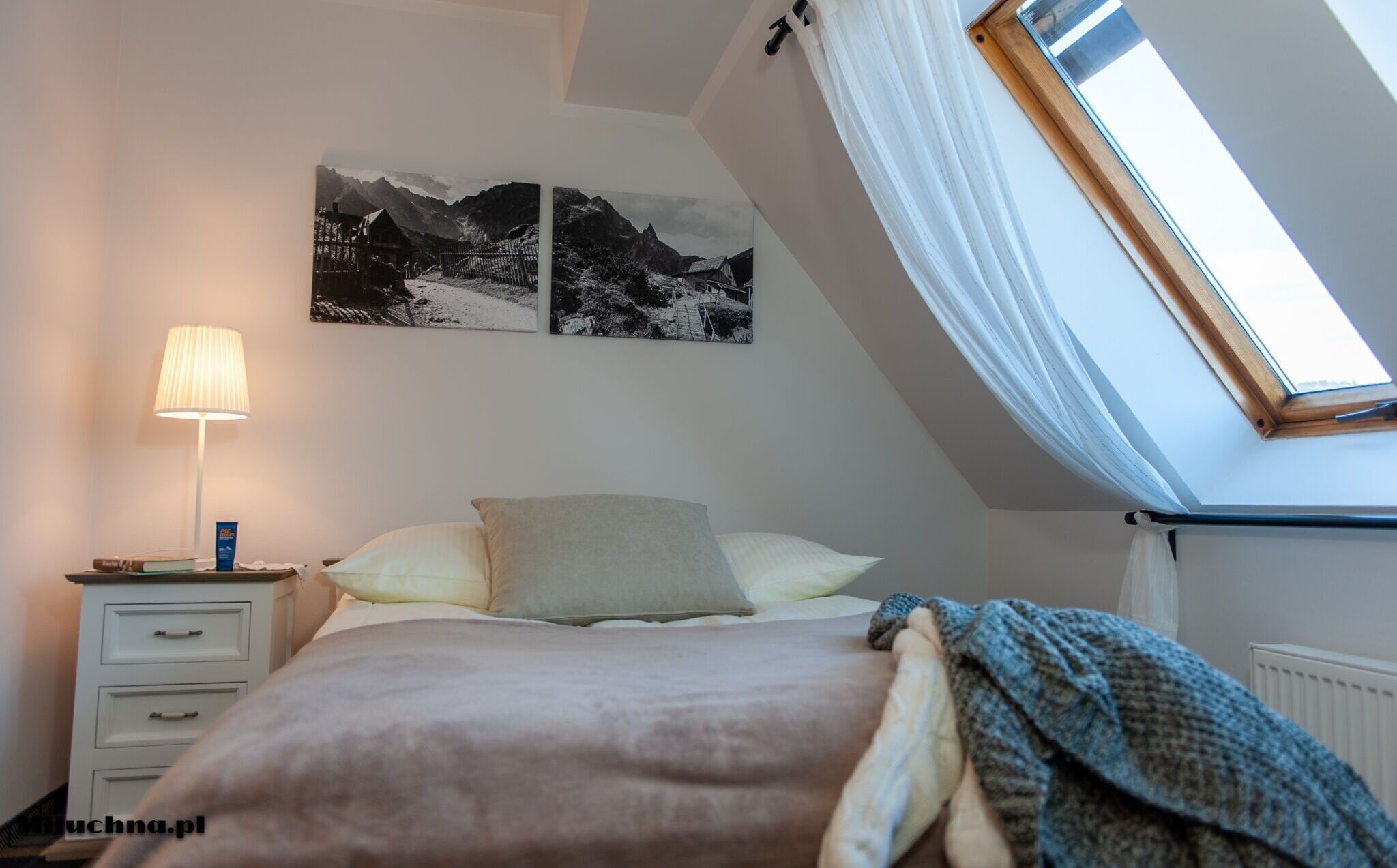 Un dormitorio con cama, mesita de noche, lámpara, ventana con vistas a montañas y dos cuadros colgados en la pared.