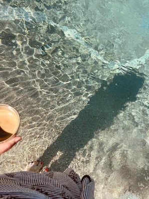 Iemand staat aan de rand van een helder, ondiep water, mogelijk een strand, met een beker in de hand.