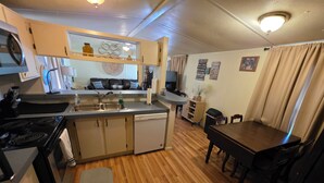 A compact kitchen with a stove, sink, and dishwasher, a dining area with a table and chairs, and a living space with a TV and decorative items. - Relax in this 2-bedroom lakefront home. Shared dock. Kayaks. Pet friendly (Cadiz)