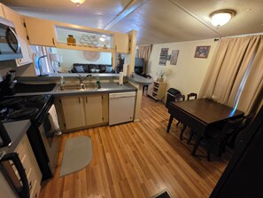 A compact kitchen with a stove, sink, and dishwasher, a dining area with a table and chairs, and a living space with a TV and decorative items. - Relax in this 2-bedroom lakefront home. Shared dock. Kayaks. Pet friendly (Cadiz)