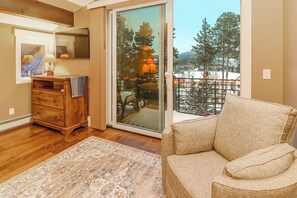 Ein Wohnzimmer mit einem Holzkleiderschrank, einem gemusterten Teppich, einem beigefarbenen Sofa und einer großen Schiebeglastür, die auf einen Balkon mit Blick auf Bäume und eine verschneite Landschaft führt.