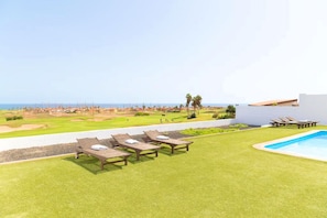 Un área de piscina con sillones de descanso, vista a un campo de golf y cielo despejado.