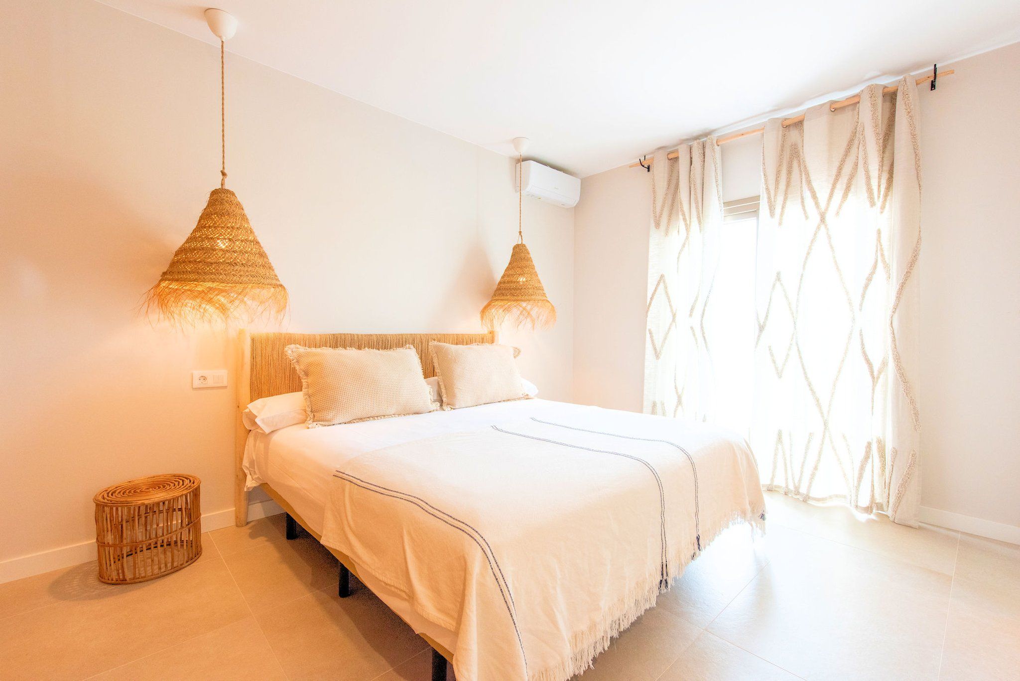 Een slaapkamer met een bed, twee hanglampen, een raam met gordijnen en een kleine rieten mand.