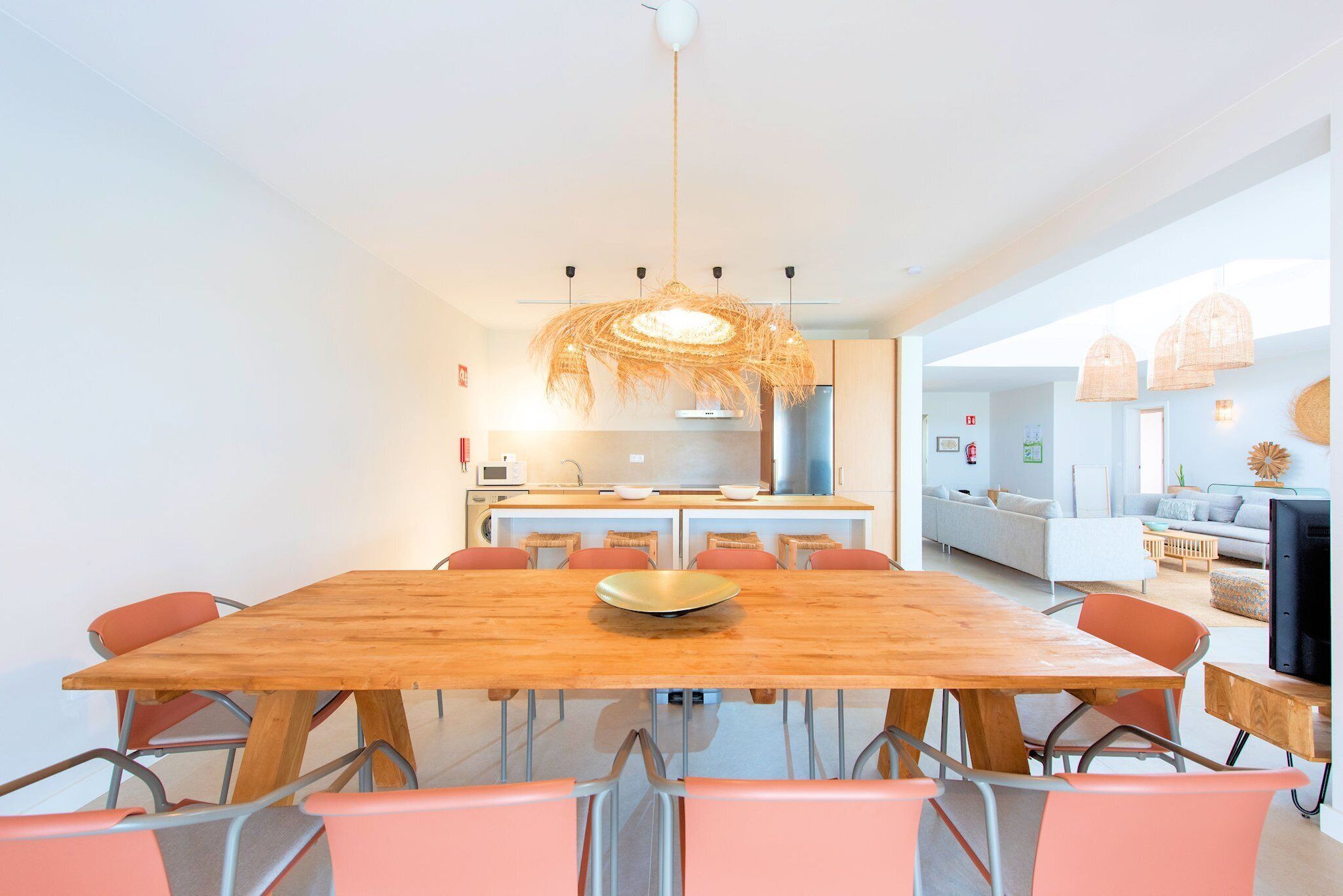 A modern dining area with a wooden table and chairs, a hanging light fixture, and a kitchen in the background.