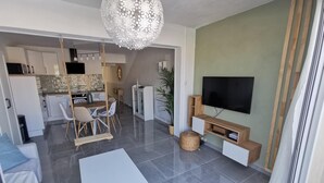 A modern living room with a flat-screen TV, a dining area with chairs, a kitchen with cabinets, and a hanging light fixture.