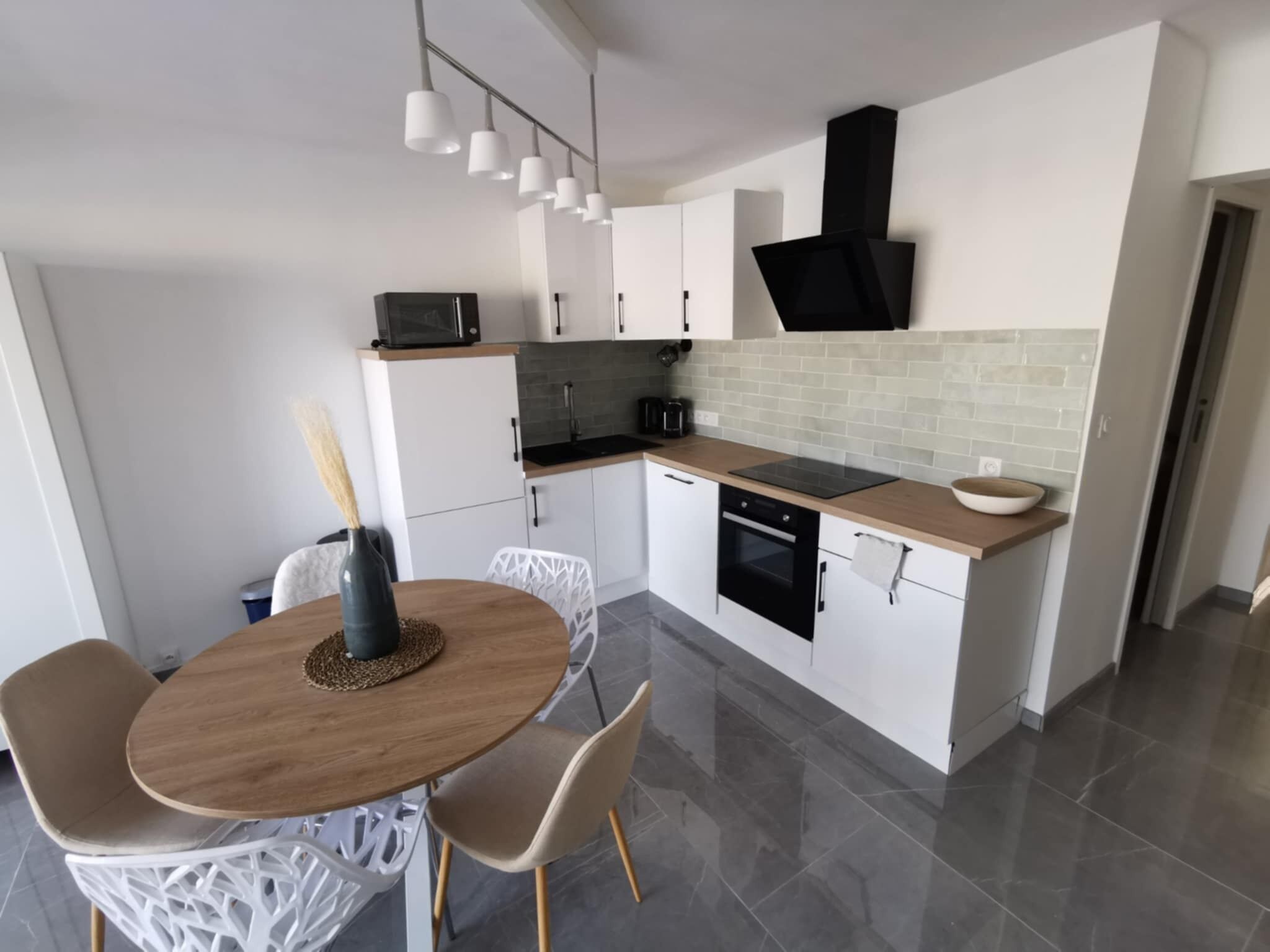 Une cuisine moderne avec des meubles blancs, une hotte noire, un four à micro-ondes et un évier. On y trouve une table à manger avec des chaises et un vase rempli de fleurs.