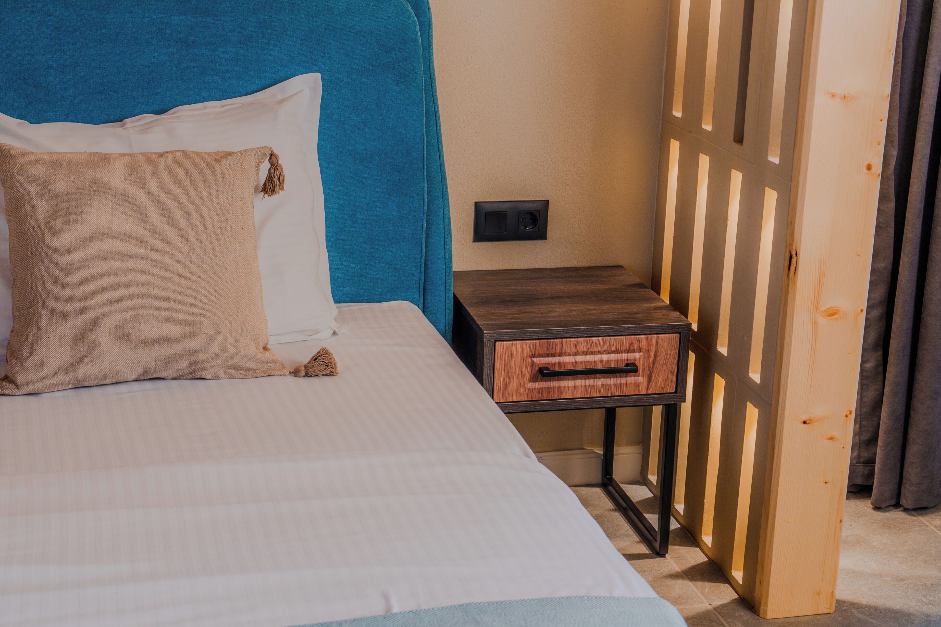 A bed with a blue headboard, a wooden nightstand with a drawer, and a wall-mounted light switch.