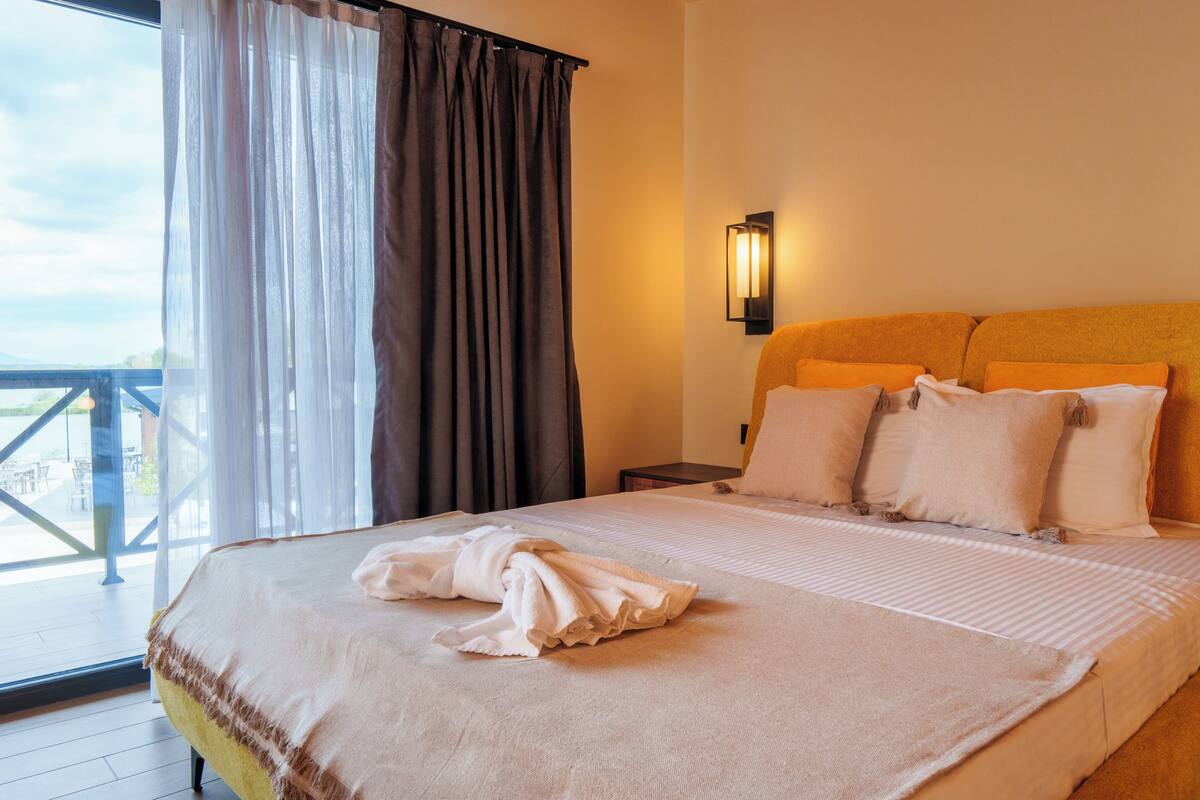 Ein Hotelzimmer mit einem Bett, einem Nachttisch, einer Wandleuchte und einem Blick durch ein Fenster mit Vorhängen.