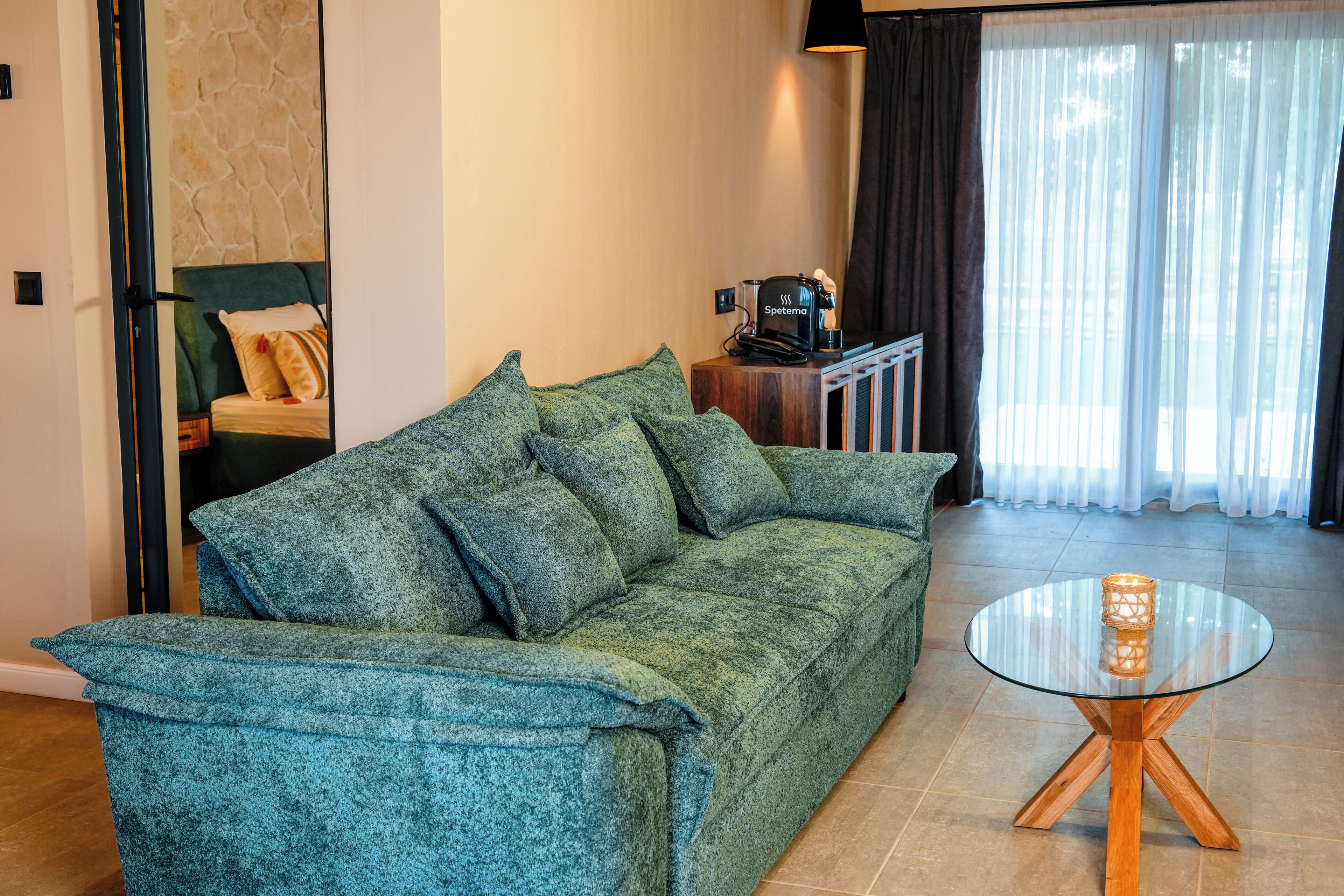 Ein grünes Sofa, ein Glastisch mit Kerze und eine Kaffeemaschine in einem Zimmer mit Steinwand und einem fenster mit Vorhängen.