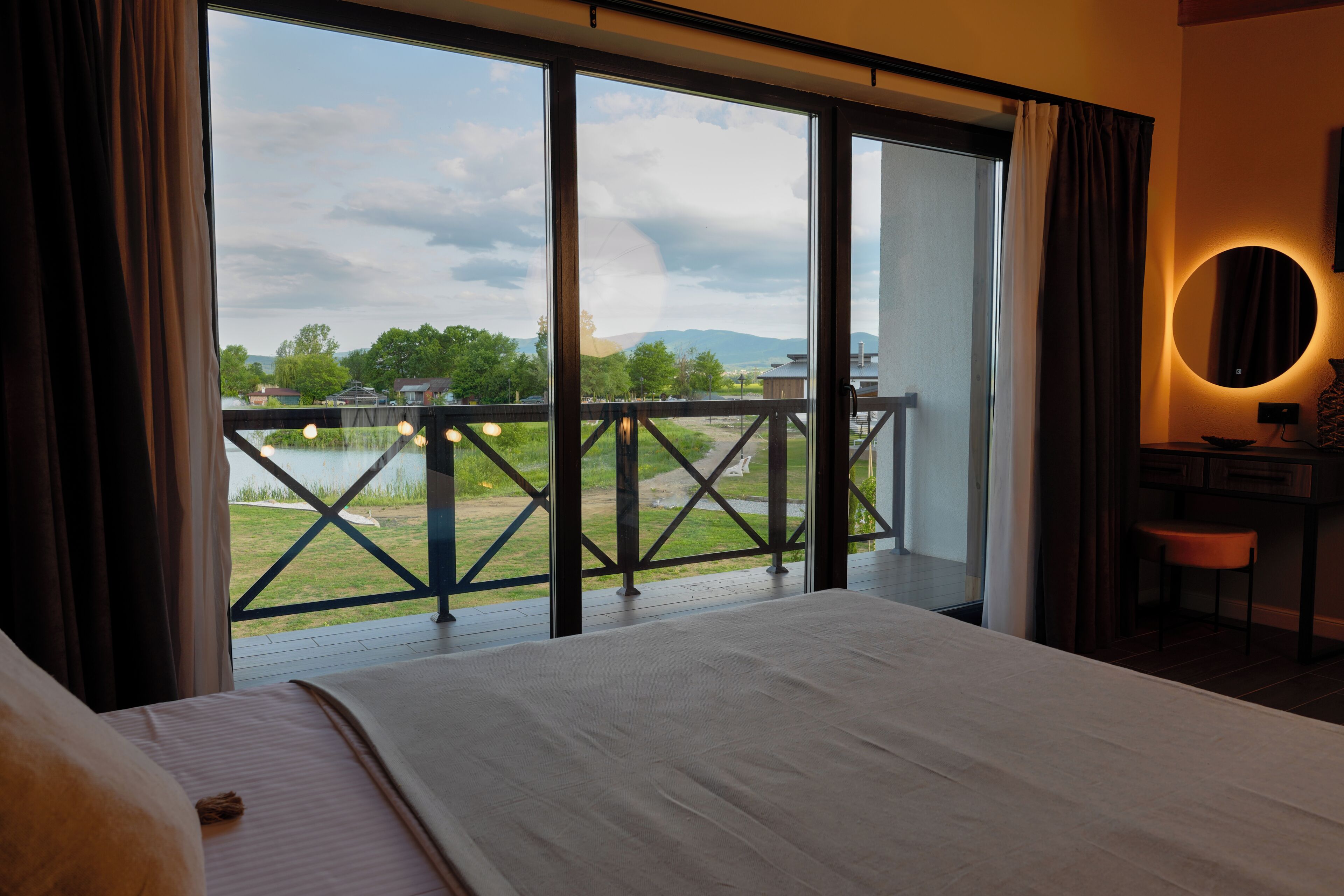 A hotel room with a large bed, a desk with a chair, a round mirror, and a balcony with a view of a lake and mountains.