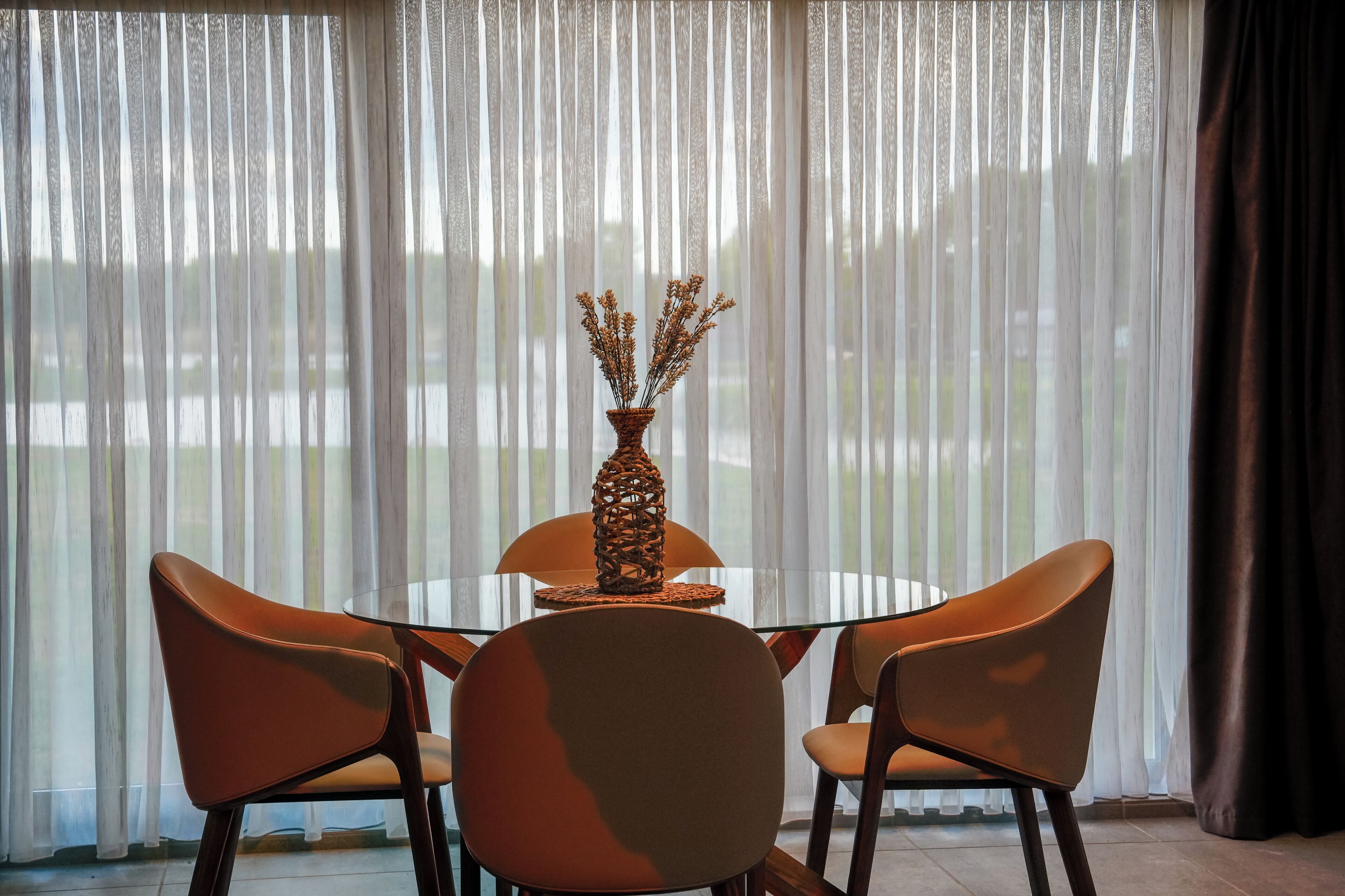 Un comedor con una mesa y sillas, un florero con plantas secas y cortinas de gasa.