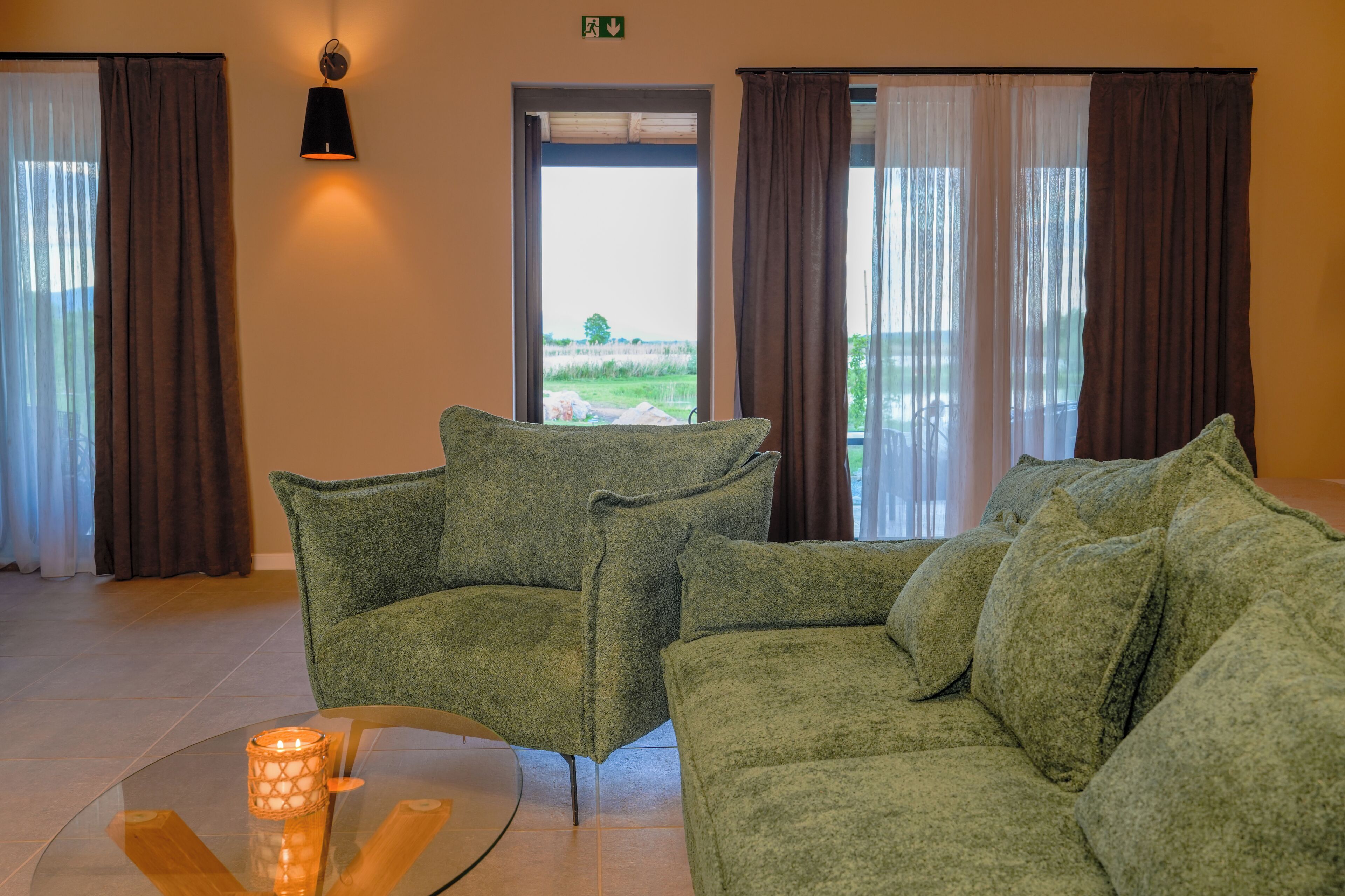 A green sofa set in a room with a glass table, a wall-mounted lamp, and a view through a door.