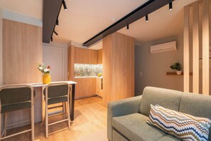 Uma sala de estar moderna com um sofá, uma mesinha com cadeiras, uma área de cozinha ao fundo e um teto com iluminação em trilhos.