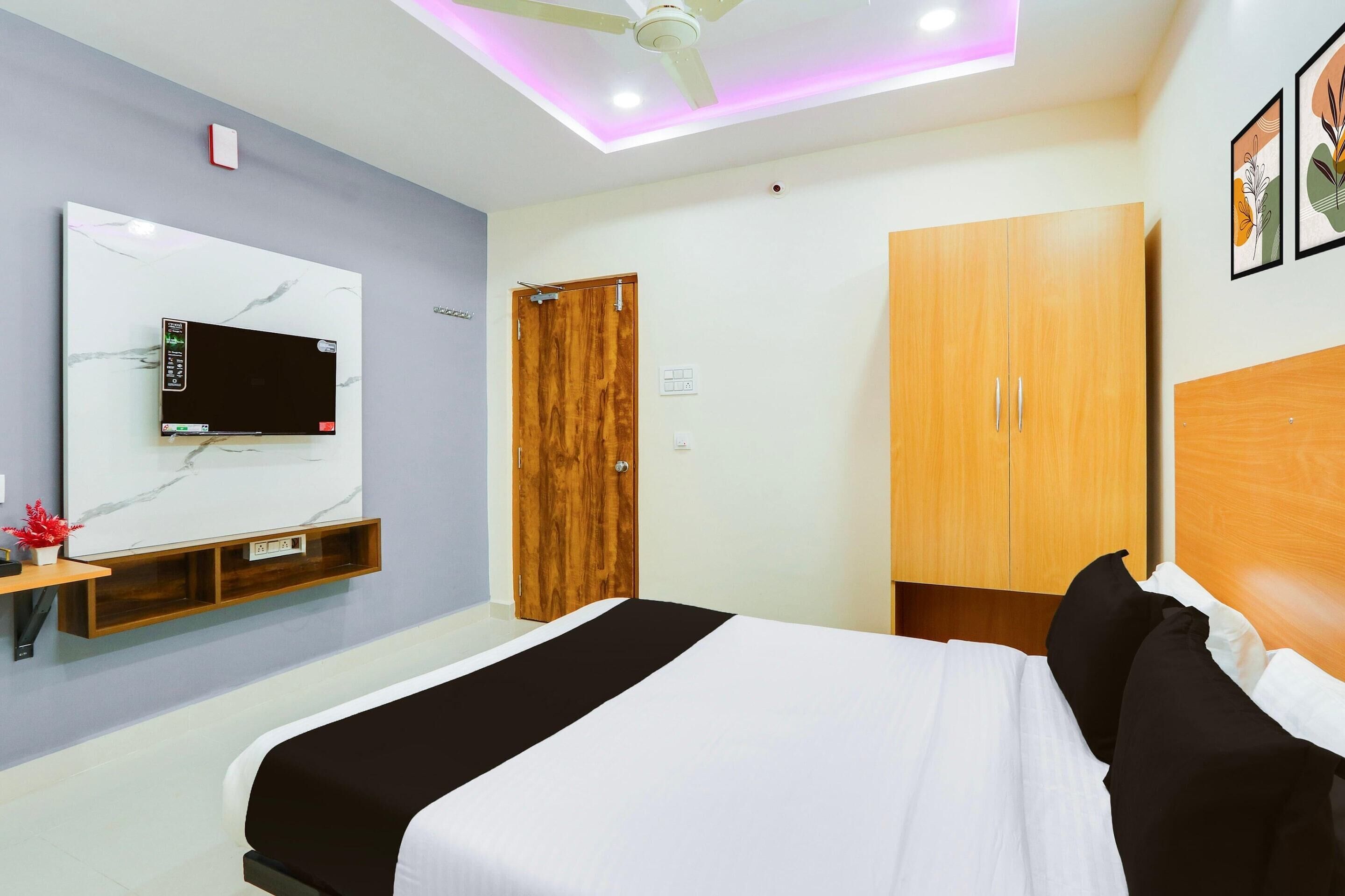 A bedroom with a bed, a TV mounted on the wall, a wooden wardrobe, and a ceiling fan.