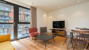 Una sala moderna con un sofá, una silla roja, una mesa de comedor con sillas, un televisor y un ventanal con vistas a edificios.