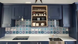 A kitchen with dark blue cabinets, a tiled backsplash, and a wooden shelf with dishes and flowers. - A little slice of heaven! (Rosmuc)