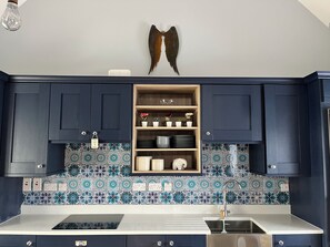 Eine Küche mit dunkelblauen Schränken, einer gefliesten Spritzschutzleiste und einem Holzregal mit Geschirr und Blumen.