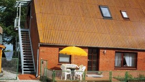 A two-story building with a metal roof, a staircase on the left, and a patio with a table and chairs. - Ferienwohnung in Orth auf Fehmarn (Fehmarn)