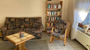 A living room with a patterned sofa, a wooden bookshelf, a small table with a vase, and a chair with a patterned cushion. - Ferienwohnung in Orth auf Fehmarn (Fehmarn)