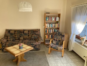 A living room with a patterned sofa, a wooden bookshelf, a small table with a vase, and a chair with a patterned cushion. - Ferienwohnung in Orth auf Fehmarn (Fehmarn)
