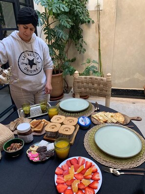 Ein Tisch ist reichhaltig mit Frühstücksleckereien wie Brot, Gebäck und Früchten gedeckt, im Hintergrund ist eine Person mit Kapuzenpulli zu sehen.