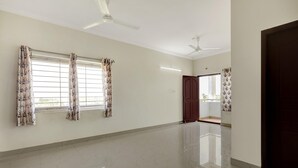 A spacious room with a polished floor, two windows with floral curtains, a ceiling fan, and a wooden door. - Classic With Balcony The Best Inn (Kakinada)
