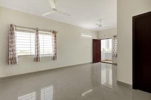 A spacious room with a polished floor, two windows with floral curtains, a ceiling fan, and a wooden door.