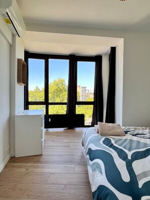 A modern bedroom with a large window, a bed with patterned bedding, a white dresser, and a built-in air conditioning unit. - Cozy and Fully Equipped in the Center of Liege 4 Guests (Liege)