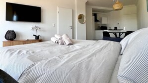 Une chambre d’hôtel comprenant un lit, une télévision, une petite table avec un vase et une plante, un coin cuisine et une table à manger.