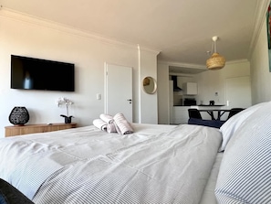 Ein Hotelzimmer mit Bett, Fernseher, kleinem Tisch mit Vase und Pflanze, Küchenzeile und Esstisch.