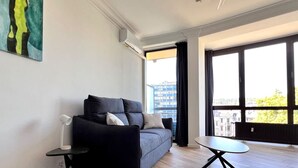 Un salon moderne avec un canapé gris, une petite table d’appoint, une table basse ronde et de grandes fenêtres donnant sur la ville.