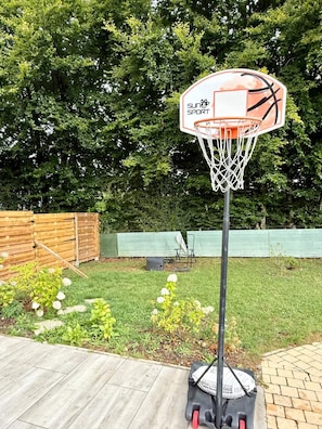 Un panier de basket avec une enseigne « SUN&SPORT », une clôture en bois et une cour verdoyante parsemée de fleurs.