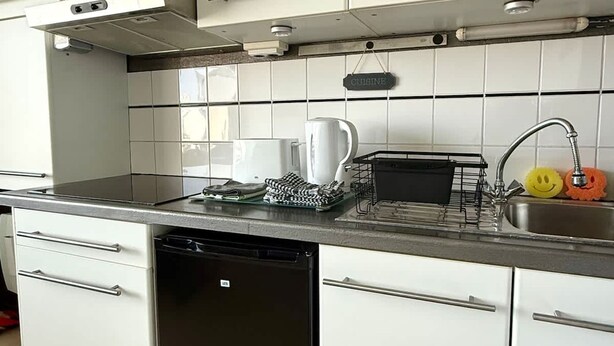 A kitchen with white cabinets, a dishwasher, a coffee maker, and a sink with a smiley face sponge. - Small Cozy Nest for 4 People 100 m From the Beach Free Parking (De Haan)