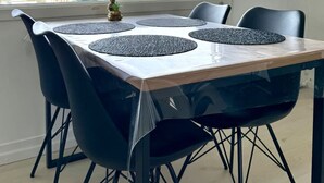 A dining area with a table, four chairs, a window with a view of buildings and trees, and a small plant on the windowsill. - Small Cozy Nest for 4 People 100 m From the Beach Free Parking (De Haan)