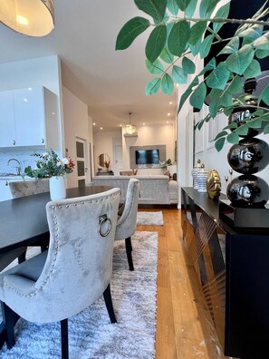 Une cuisine moderne avec des meubles blancs, un coin repas doté d’une chaise recouverte de tissu blanc, et un salon avec un canapé gris.