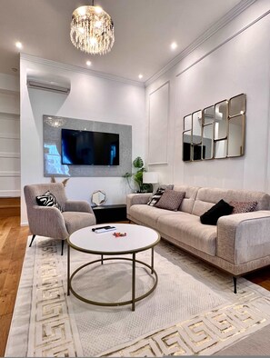 Un salon moderne avec un téléviseur à écran plat, une table basse ronde, un canapé beige et un lustre.